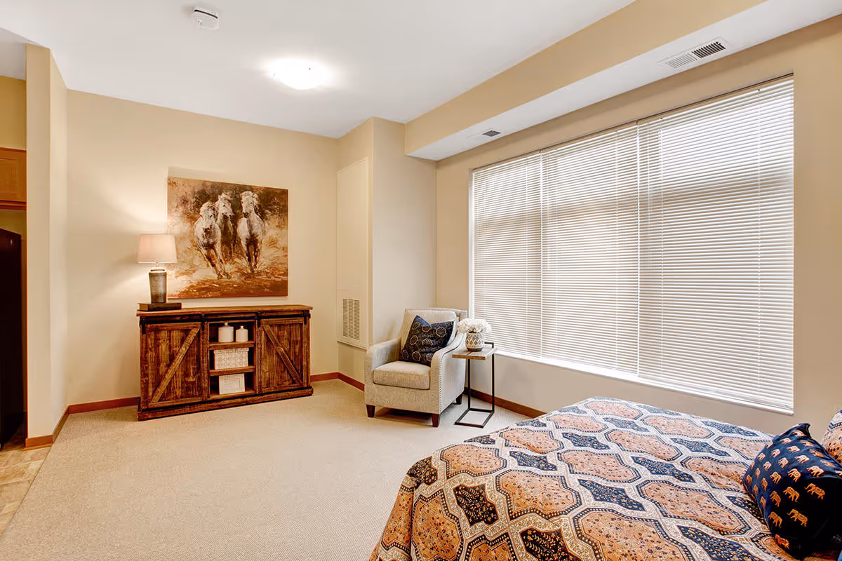 A cozy bedroom with a large window covered by blinds, a patterned bedspread on the bed, a cushioned armchair with a pillow, a small side table with a flower vase, and a wooden cabinet with a lamp and a painting of running horses above it.