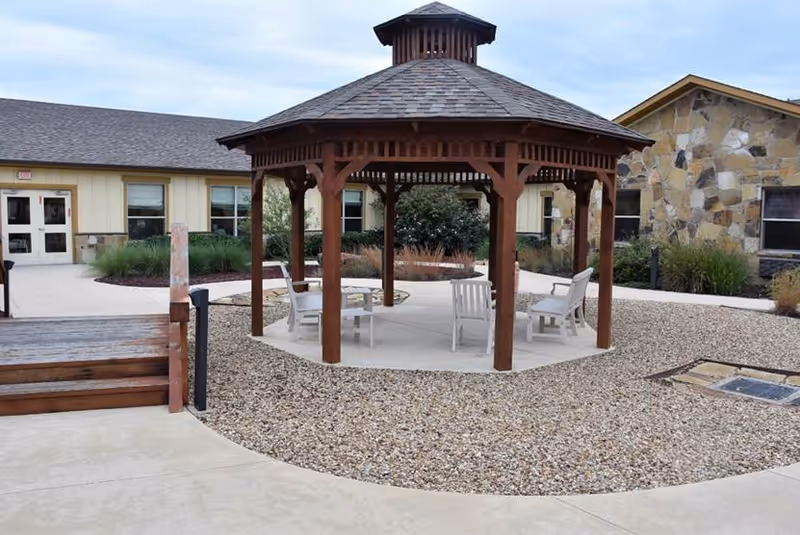 Outdoor courtyard area featuring a wooden gazebo with a shingled roof, surrounded by four white chairs on a concrete pad. The ground around the gazebo is covered with small gravel stones. In the background, there are buildings with beige and stone exterior walls and several windows. There is also a wooden ramp and concrete walkways leading to the buildings.