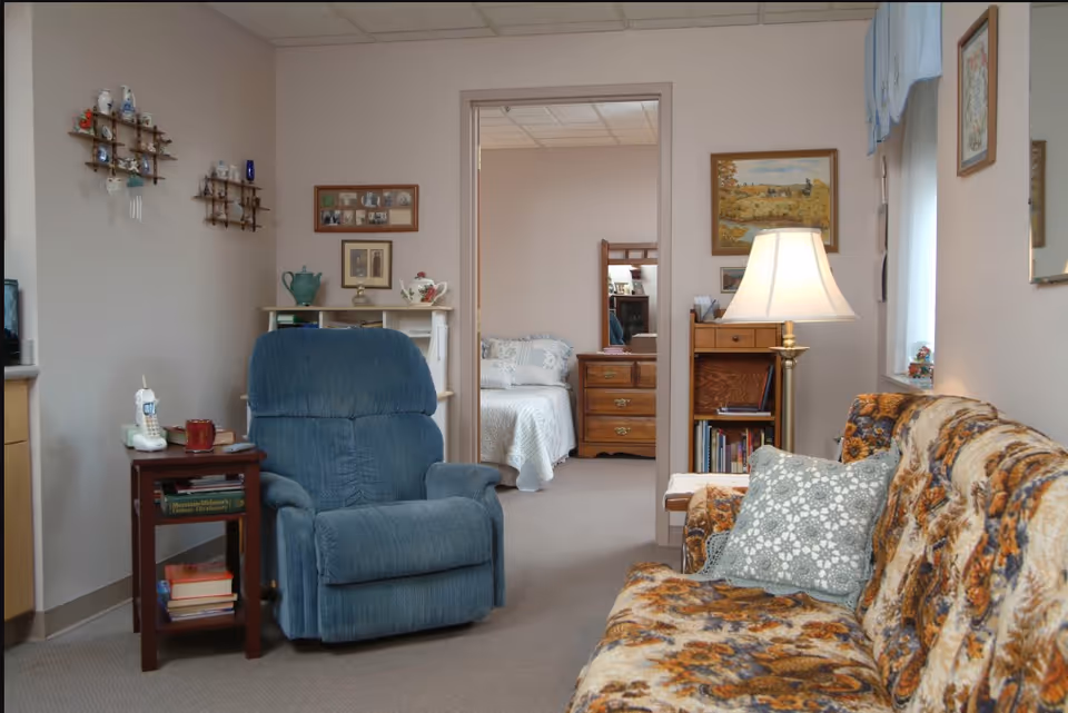 A cozy living room area with a floral patterned sofa and a blue recliner chair. There is a wooden side table with a cordless phone and books next to the recliner. A lamp on a small wooden cabinet illuminates the room. The walls are decorated with framed pictures and small shelves holding decorative items. Through an open doorway, a bedroom with a bed and wooden dresser is visible.