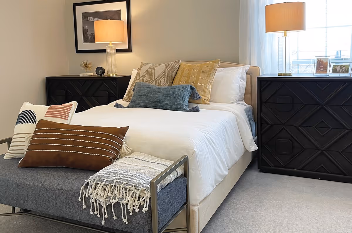A neatly made bed with white bedding and multiple decorative pillows in various colors including blue, brown, and beige. At the foot of the bed is a gray bench with a fringed throw blanket and additional pillows. On either side of the bed are black nightstands with geometric patterns, each topped with a lamp with a beige shade. A framed black and white photo hangs on the wall above the left nightstand, and the right nightstand has two framed photos. A window with sheer white curtains is visible on the right side of the room.