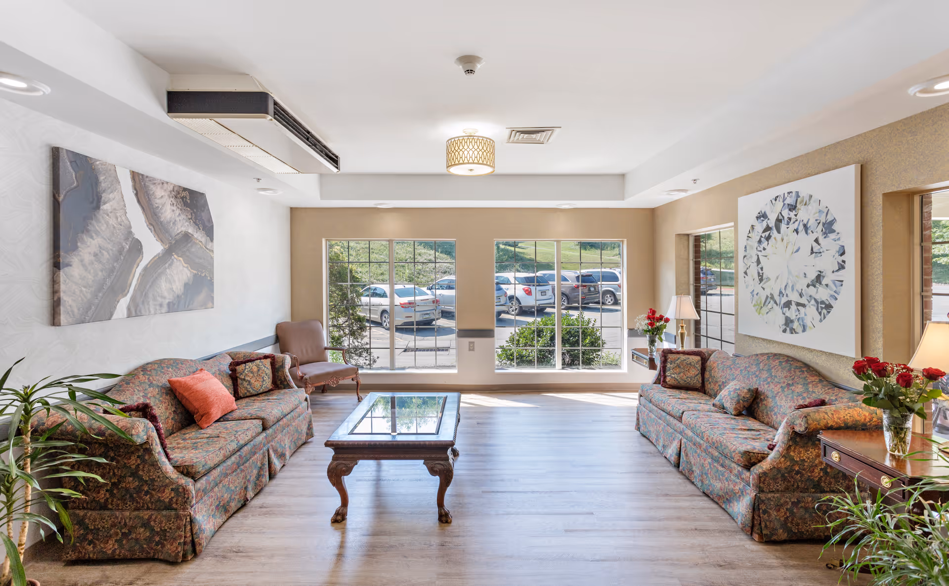 A bright lounge with two patterned sofas, a glass-top coffee table, potted plants, and large windows overlooking a parking lot.