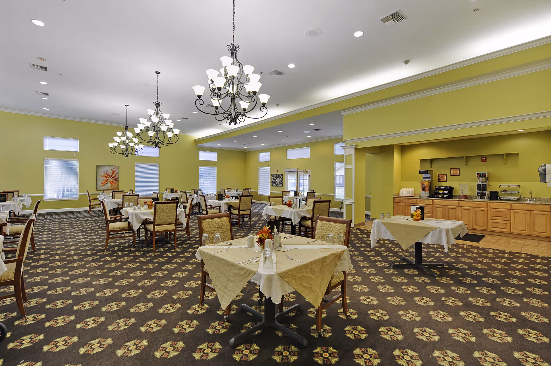 Spacious dining room with multiple cloth-covered tables and chairs, ornate chandeliers, patterned carpet, and a serving counter along the right wall.