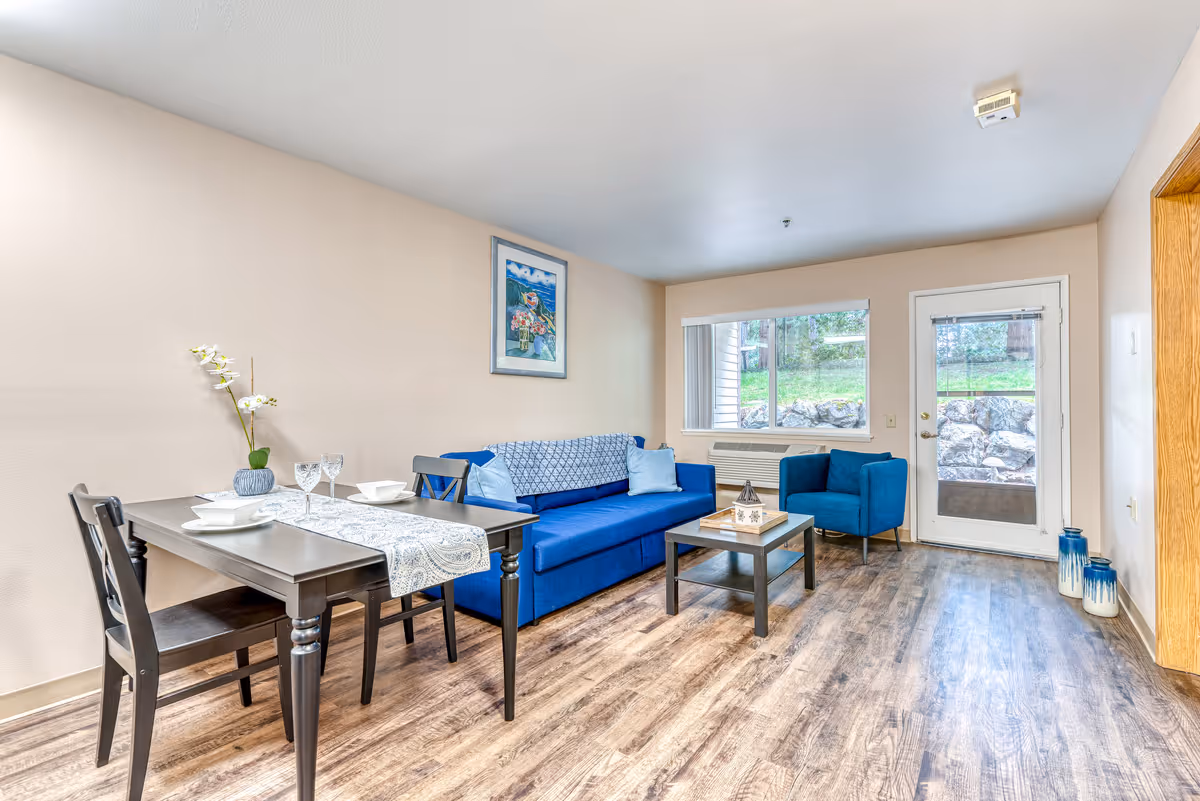 A bright living room and dining nook with a blue sofa and armchair, coffee table, small dining table set, and a glass door and window overlooking a rocky yard.