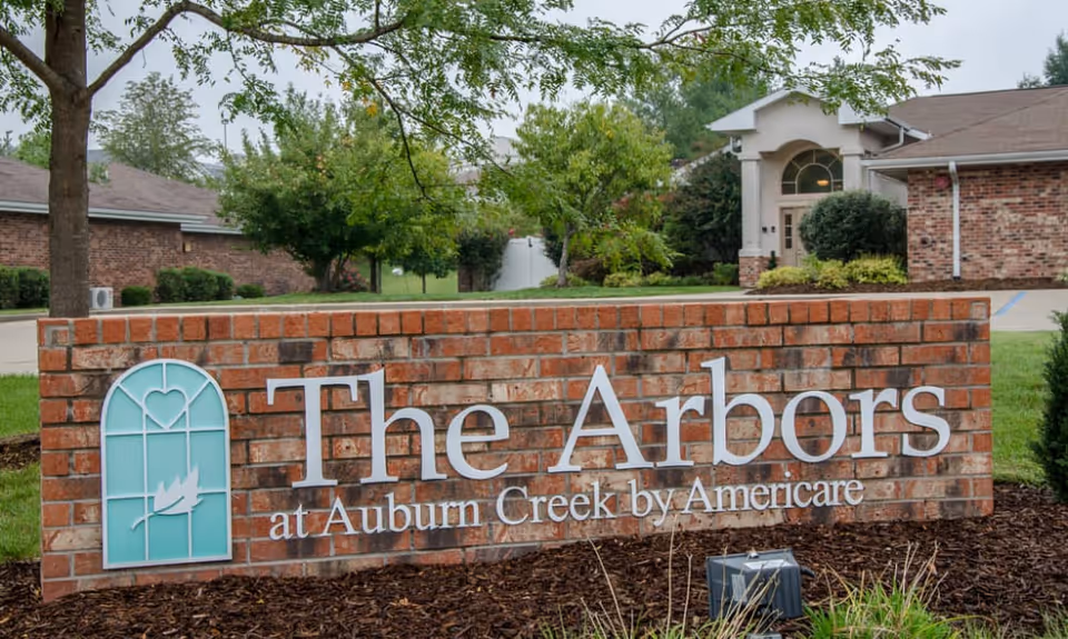 A brick sign for The Arbors at Auburn Creek by Americare in front of a senior living facility building with trees and landscaping around.