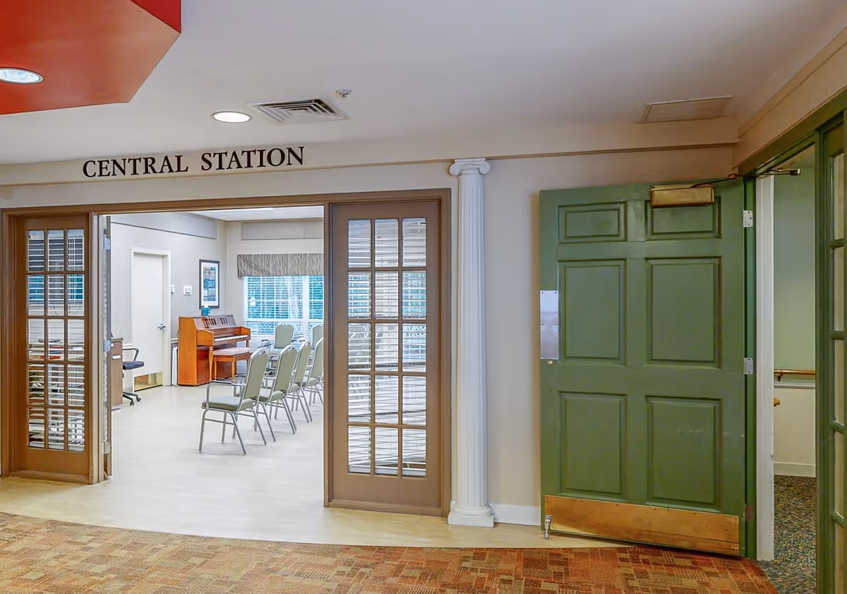 Interior view of a room labeled 'Central Station' with glass-paneled double doors open to a room containing several chairs arranged in rows and a piano against the far wall. The area outside the room has a green door and a white decorative column.