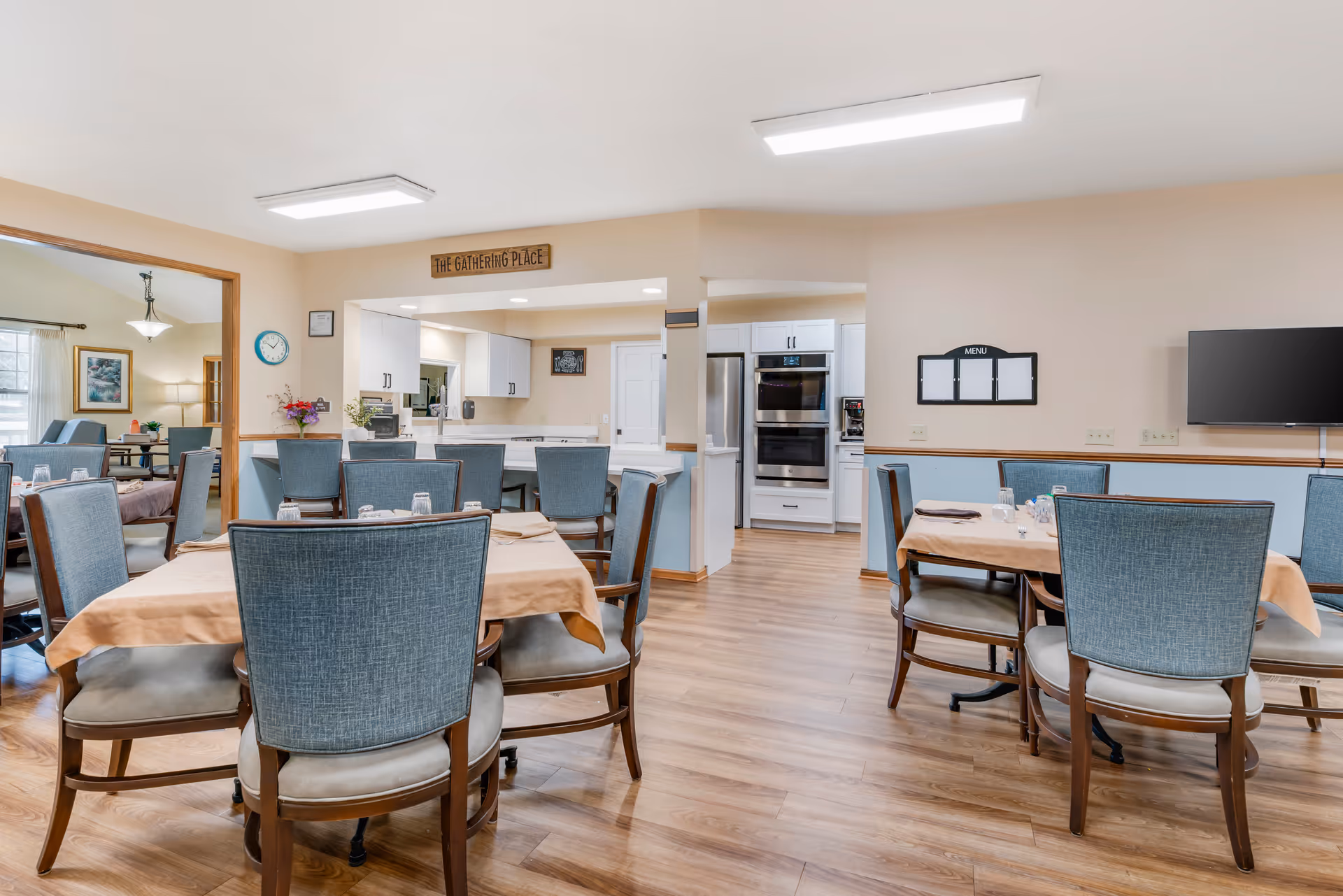 A dining area in a senior living facility with several tables covered with beige tablecloths and surrounded by blue upholstered chairs. The room has wood flooring and beige walls with a light blue wainscoting. In the background, there is a kitchen area with white cabinets, stainless steel appliances, and a sign above the kitchen pass-through window that reads 'The Gathering Place'. A wall-mounted TV and a blank menu board are visible on the right wall.