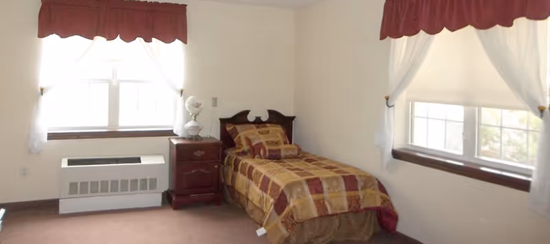 A small bedroom with a single bed covered in a patterned bedspread in shades of gold and burgundy. There is a wooden nightstand next to the bed with a decorative lamp on it. Two windows with white curtains and burgundy valances let in natural light. The walls are light-colored and the floor is carpeted.