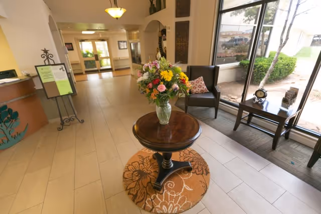 Reception lobby with a round table holding a vase of flowers, chairs by large windows, and a front desk in the background.
