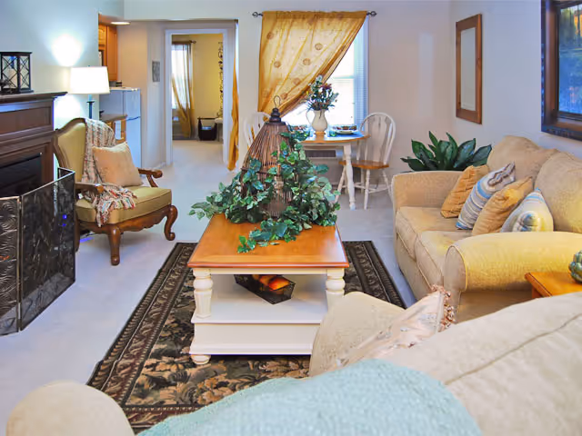 A cozy living room with beige sofas adorned with patterned pillows, a wooden coffee table with a decorative plant centerpiece, an armchair with a throw blanket, a fireplace on the left, and a small dining table with two chairs near a window with a yellow curtain.