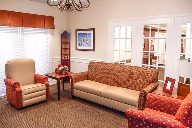 A cozy living room area with a patterned brown sofa, two red patterned armchairs, a small wooden side table with a basket of fruit, a framed picture on the wall, and a chandelier overhead. The room has beige walls, a carpeted floor, and white French doors leading to another room.