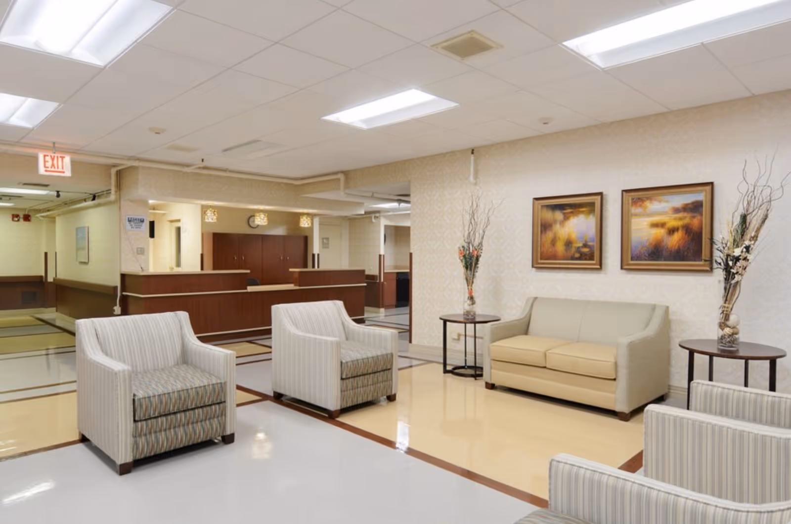 A clean and well-lit waiting area in a senior living facility with beige walls and tiled floors. The room features a beige sofa, two striped armchairs, two round side tables with decorative vases holding dried branches, and two framed landscape paintings on the wall. In the background, there is a reception desk with wood paneling and overhead lighting, and an exit sign is visible on the ceiling.