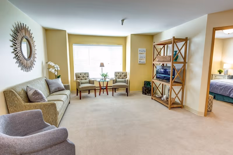 A bright living room in a senior living facility with a beige sofa, two armchairs around a small round table by a large window, a wooden shelving unit with a TV, and a glimpse into a bedroom with a bed and nightstand with a lamp.
