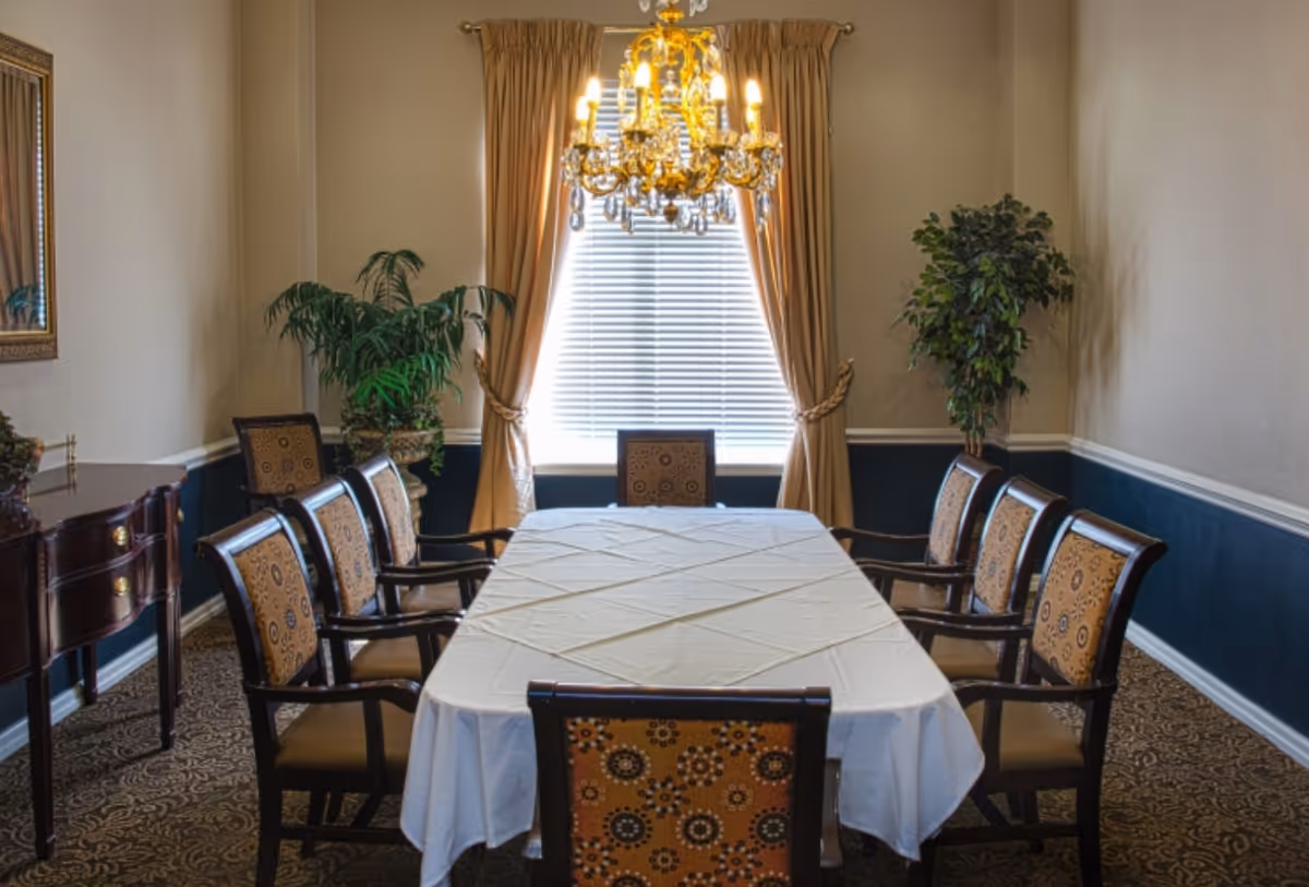A formal dining room with a rectangular table covered with a white tablecloth, surrounded by eight patterned chairs. A large window with beige curtains and blinds is centered on the back wall, illuminated by a gold chandelier with multiple lights. There are two potted plants on either side of the window and a wooden sideboard with a mirror on the left wall.