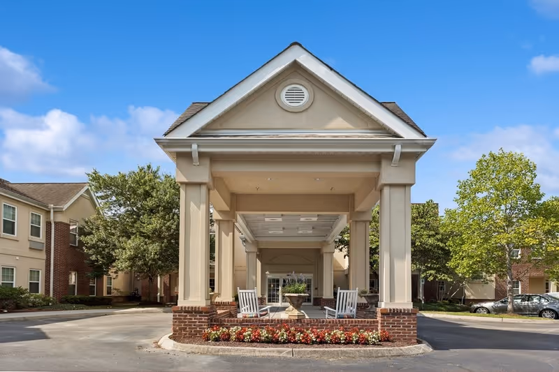 Front entrance of a senior living facility with a covered driveway supported by columns, two white rocking chairs, a planter with flowers, and surrounding trees and buildings under a blue sky.