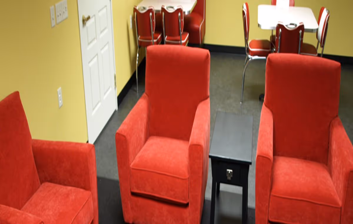 Interior room with three red upholstered armchairs arranged around a small black side table. In the background, there is a white door and a dining area with a white table and red chairs against yellow walls.