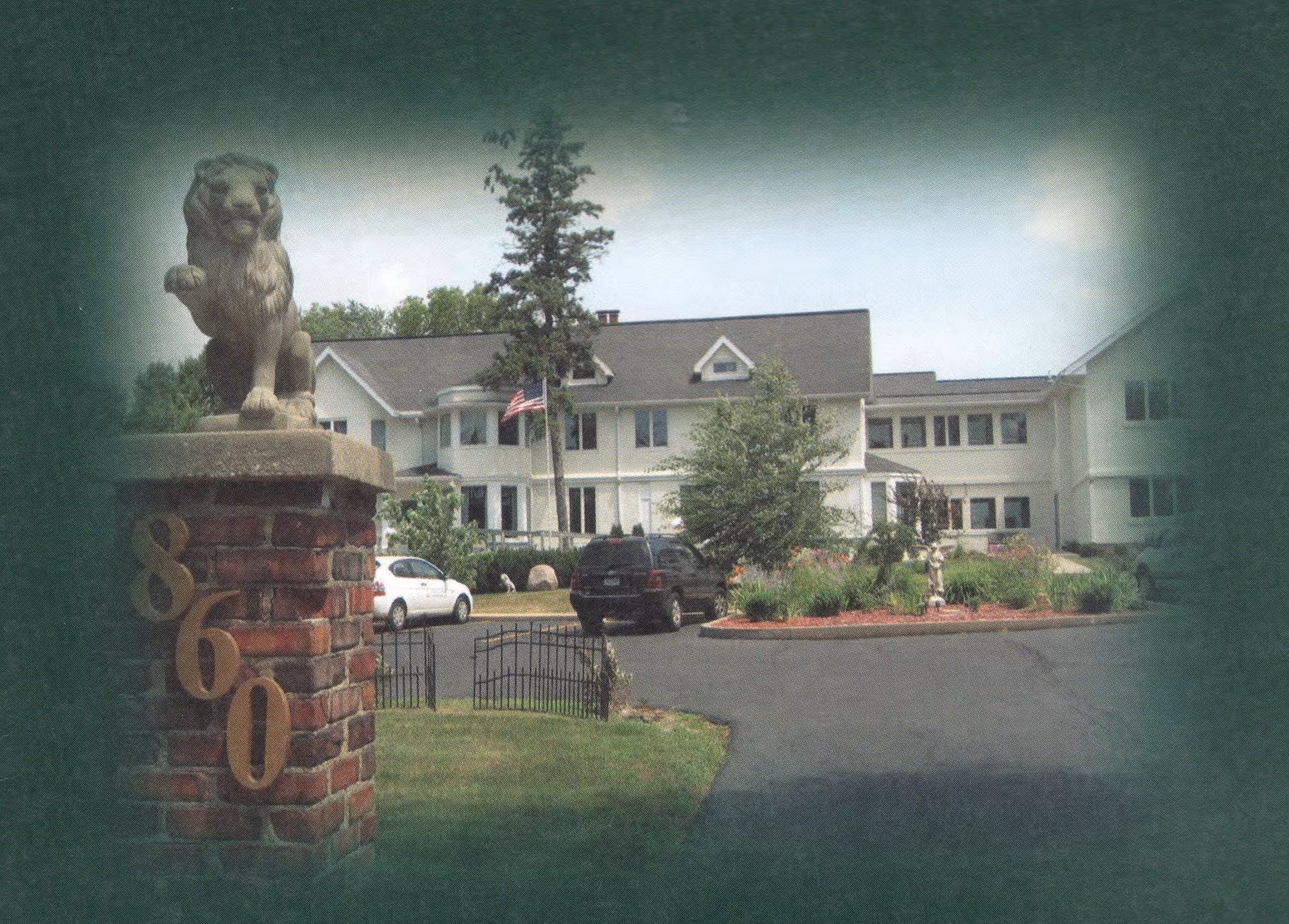 Front exterior of a multi-story care facility seen from the driveway with a lion statue on a brick pillar marked '860', American flag, parked cars, and landscaping.