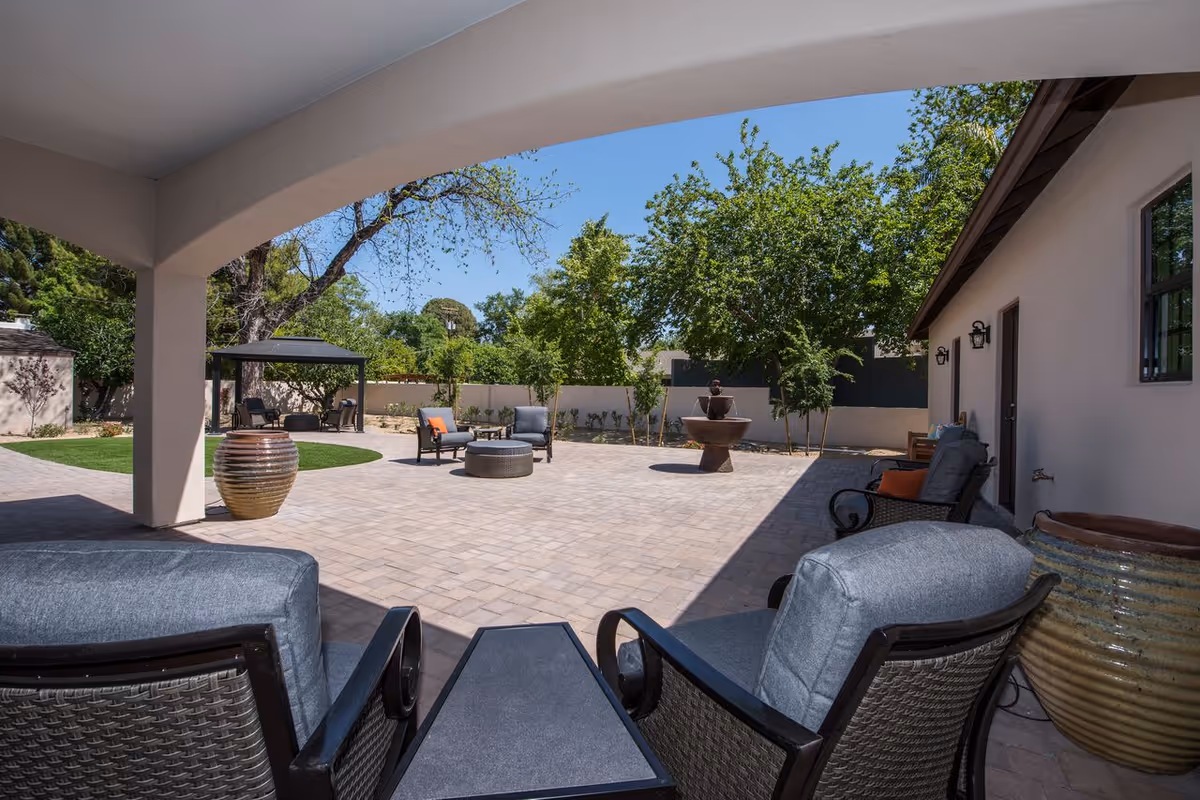 Shaded patio seating area overlooking a paved courtyard with lounge chairs, a central fountain, large planters, and trees.