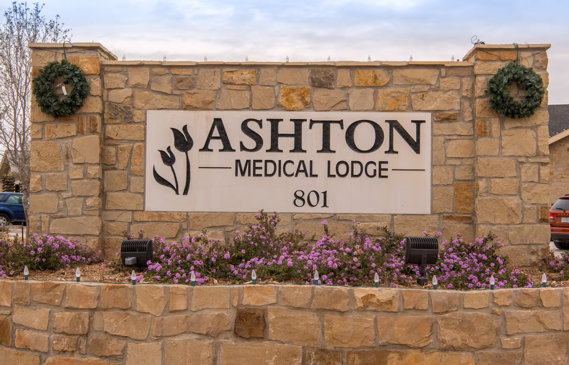 Stone sign for Ashton Medical Lodge with the number 801, decorated with two green wreaths on either side and purple flowers planted in front.