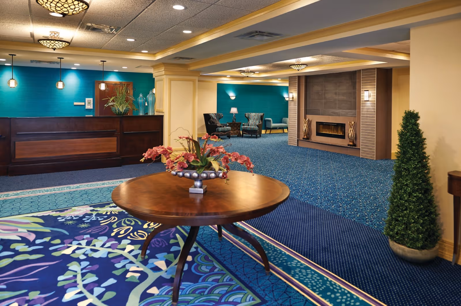 A spacious and well-lit assisted living facility lobby with a round wooden table in the foreground holding a floral arrangement. The room features blue patterned carpeting, a dark wooden reception desk against a teal accent wall, and a seating area with two patterned armchairs and a small table with a lamp. There is a modern fireplace with decorative elements on either side and a tall potted plant near the right wall.