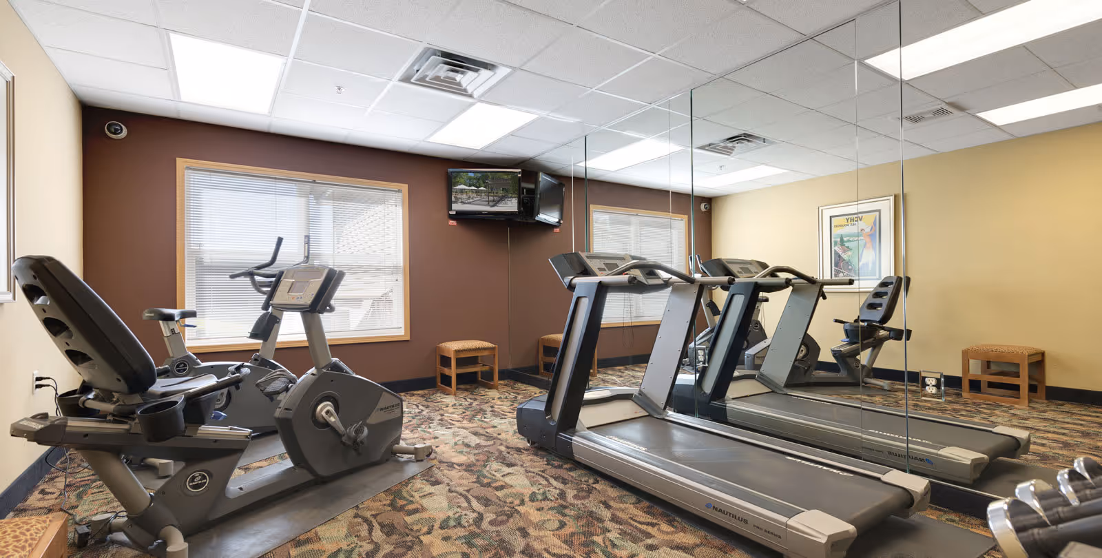Small fitness room with a treadmill, stationary bike, and recumbent bike. The room has a patterned carpet, beige and brown walls, a large mirror on one wall, two windows with blinds, a wall-mounted TV, and two small wooden stools.