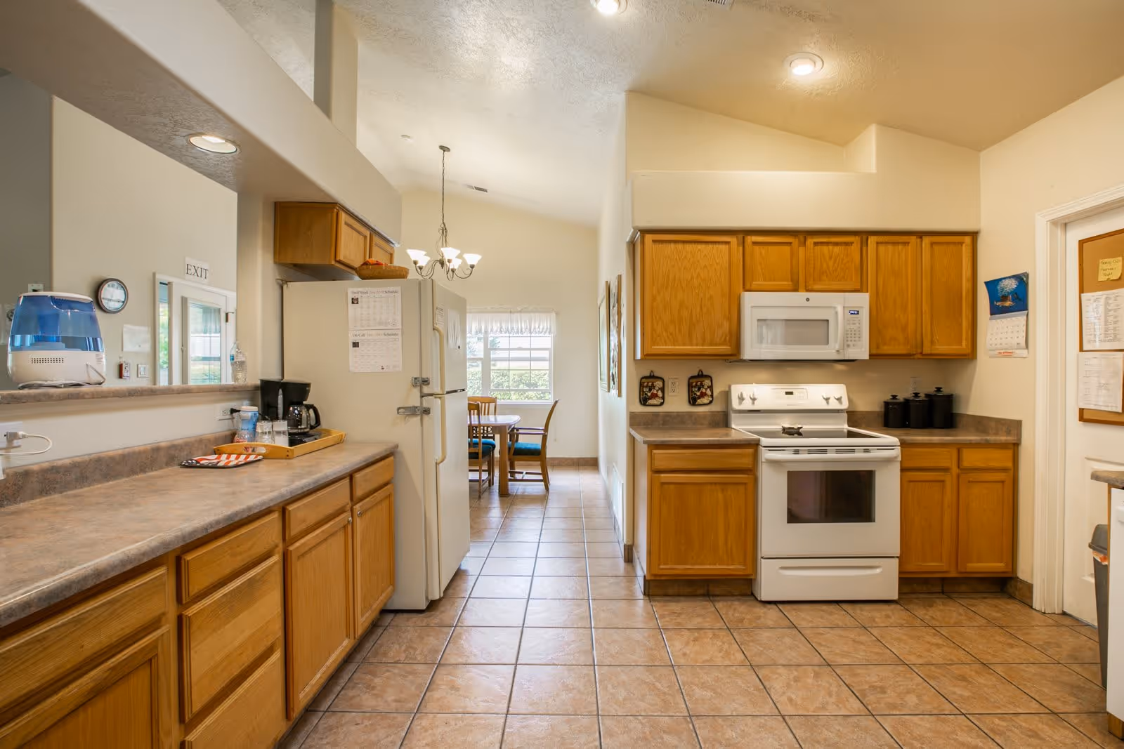A clean kitchen with wooden cabinets, a white stove and microwave, a refrigerator, and a countertop with a coffee maker and water dispenser. In the background, there is a dining area with a table and chairs near a window.
