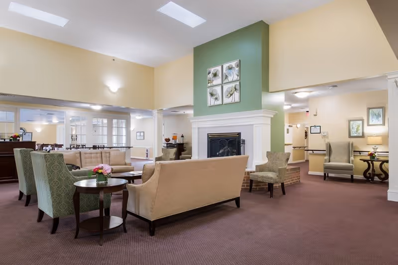 A spacious and well-lit living room area in a senior living facility featuring beige and green upholstered chairs and sofas arranged around a central fireplace with a green accent wall. The room has a carpeted floor, soft lighting, and decorative floral artwork above the fireplace. There are additional seating areas and tables visible in the background.