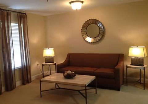 A cozy living room with a brown sofa centered against a beige wall. Above the sofa is a round decorative mirror. On either side of the sofa are small side tables, each with a lamp that has a rectangular base and a light-colored shade. A rectangular coffee table with a light wood top and dark metal legs is placed in front of the sofa, holding a decorative bowl with assorted items. To the left, there is a window with brown and beige striped curtains.