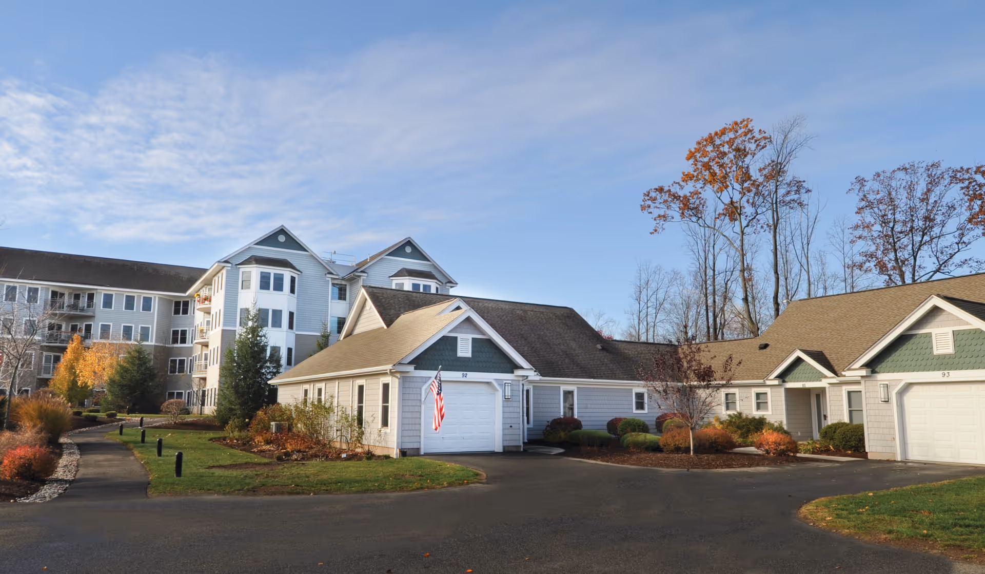 Exterior view of a senior living facility with multiple connected buildings, including a multi-story building and single-story units with garages. The area is landscaped with trees and shrubs, and a paved driveway and walkway are visible under a partly cloudy sky.