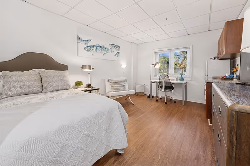 A bright and clean bedroom with a large bed covered in white bedding, two patterned pillows, a bedside table with a lamp and plant, a white cushioned chair, a desk with a chair and lamp near a window, and wooden flooring. The room has white walls and ceiling tiles, with a piece of artwork featuring fish above the bed.