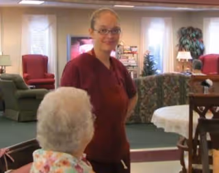 A caregiver in a maroon uniform stands and smiles in a living room area of an assisted living facility. An elderly person with white hair is seated in the foreground, facing the caregiver. The room has comfortable chairs, a table with a white tablecloth, and a television in the background.