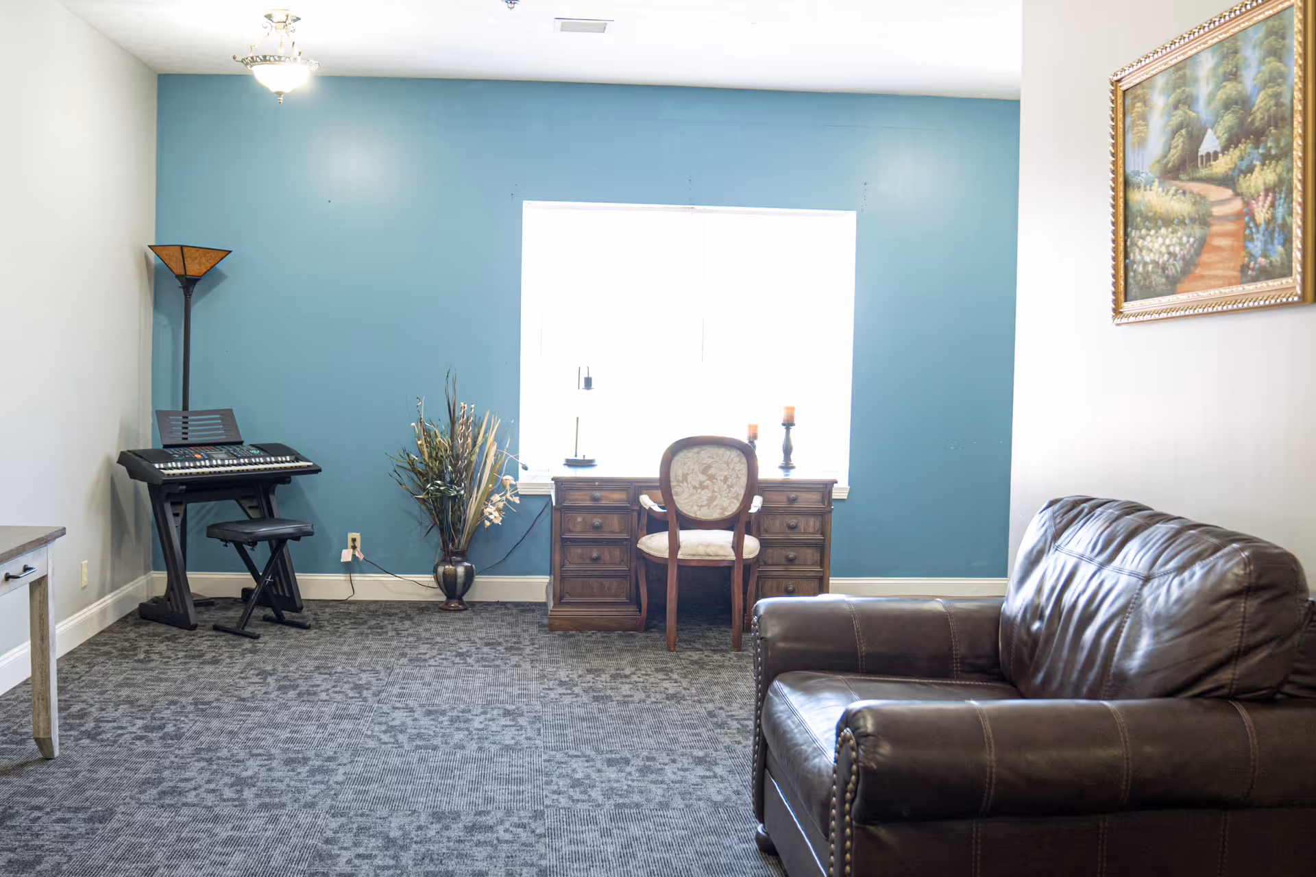 A cozy room with a blue accent wall featuring a wooden desk and chair in front of a window. To the left, there is an electronic keyboard with a stool and a tall floor lamp. A vase with decorative dried plants is placed near the desk. On the right side, there is a dark brown leather armchair and a framed painting of a garden path on the wall.