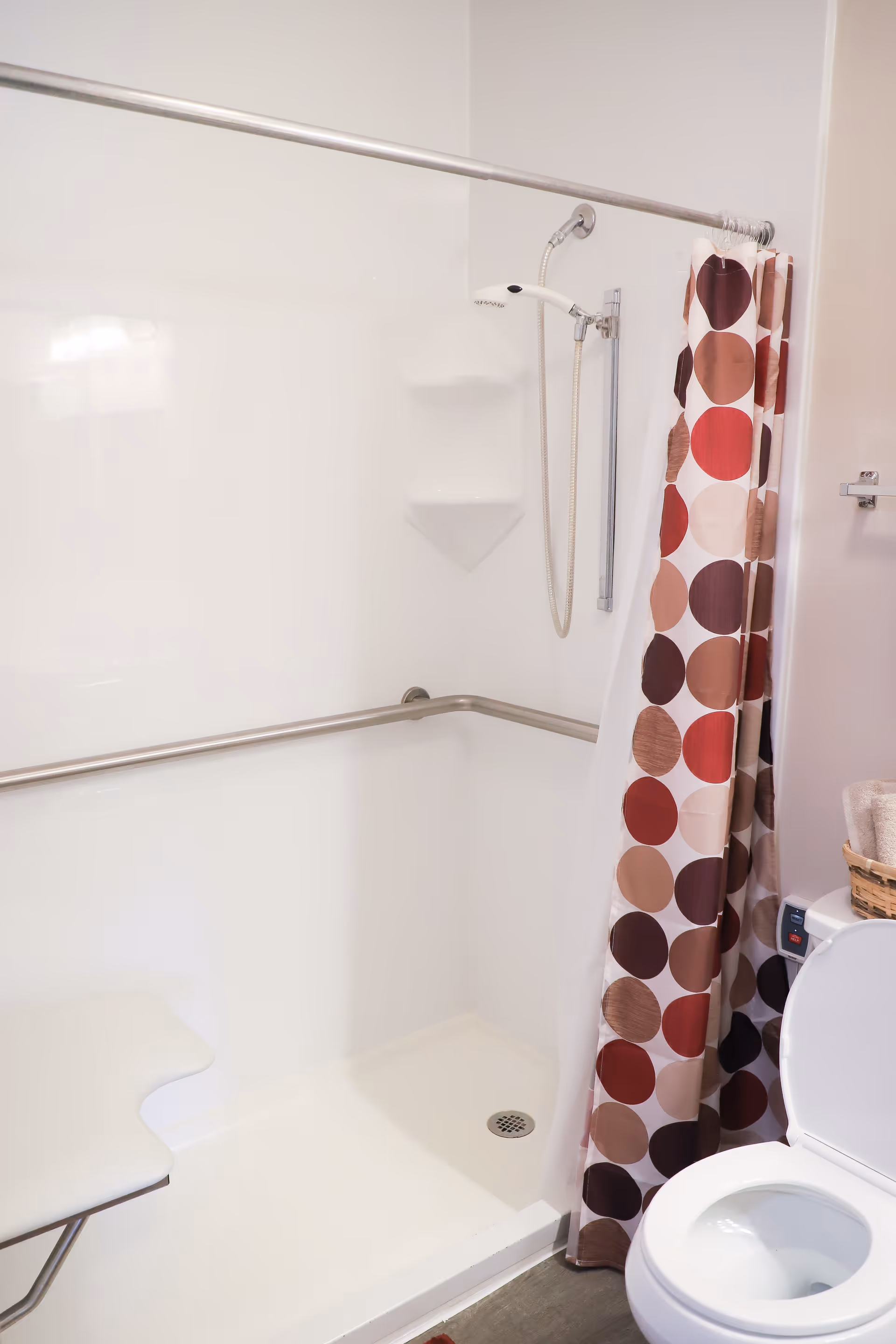 A bathroom shower area with a white shower seat, grab bars along the walls, a handheld showerhead, and a shower curtain with large circular patterns in shades of brown, red, and beige. A toilet with an open lid and a basket with towels are partially visible on the right side.