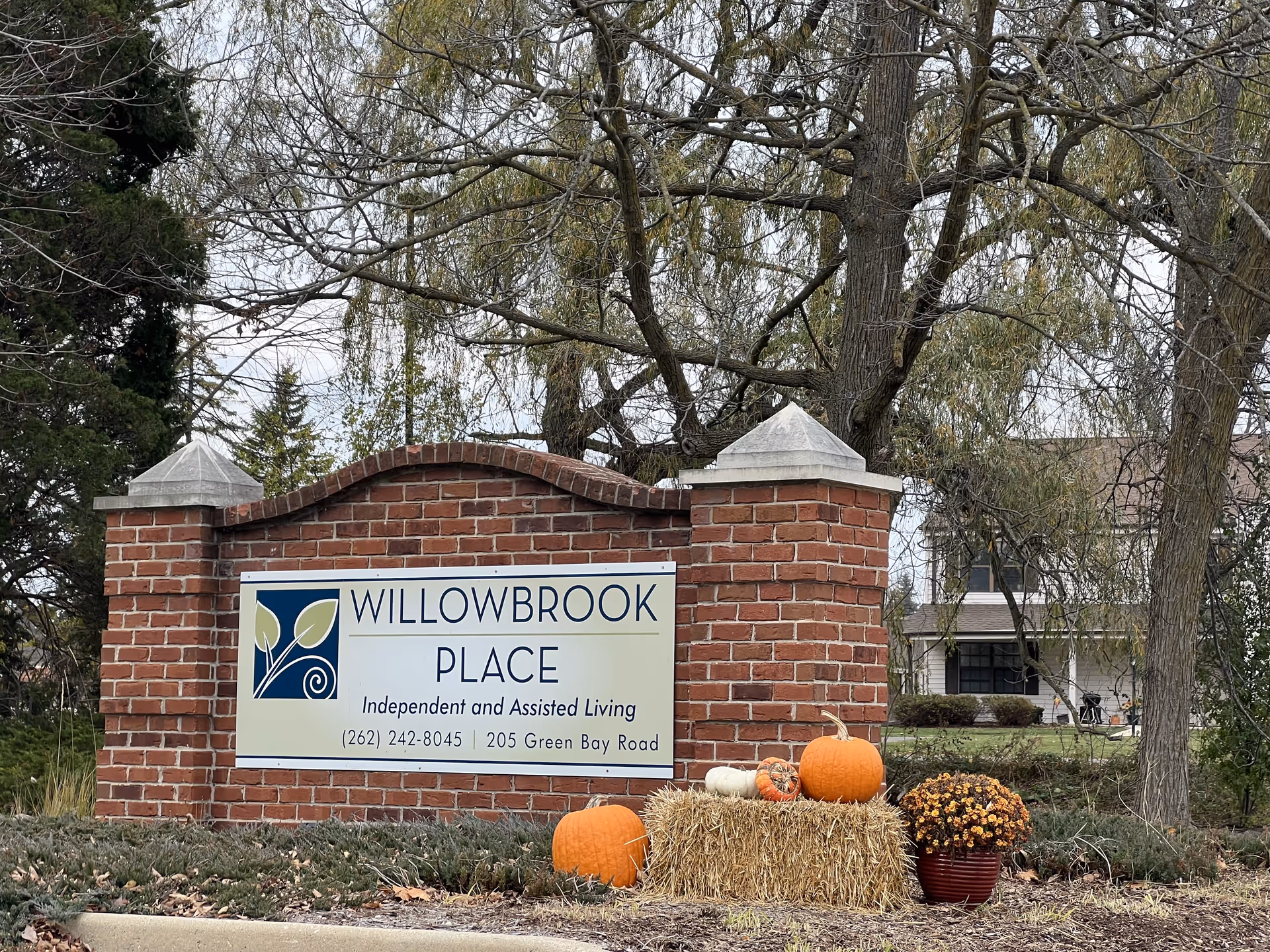 A brick sign for Willowbrook Place, an independent and assisted living facility, with the address 205 Green Bay Road and phone number (262) 242-8045. The sign is decorated with pumpkins, a hay bale, and a pot of flowers, with trees and a building visible in the background.