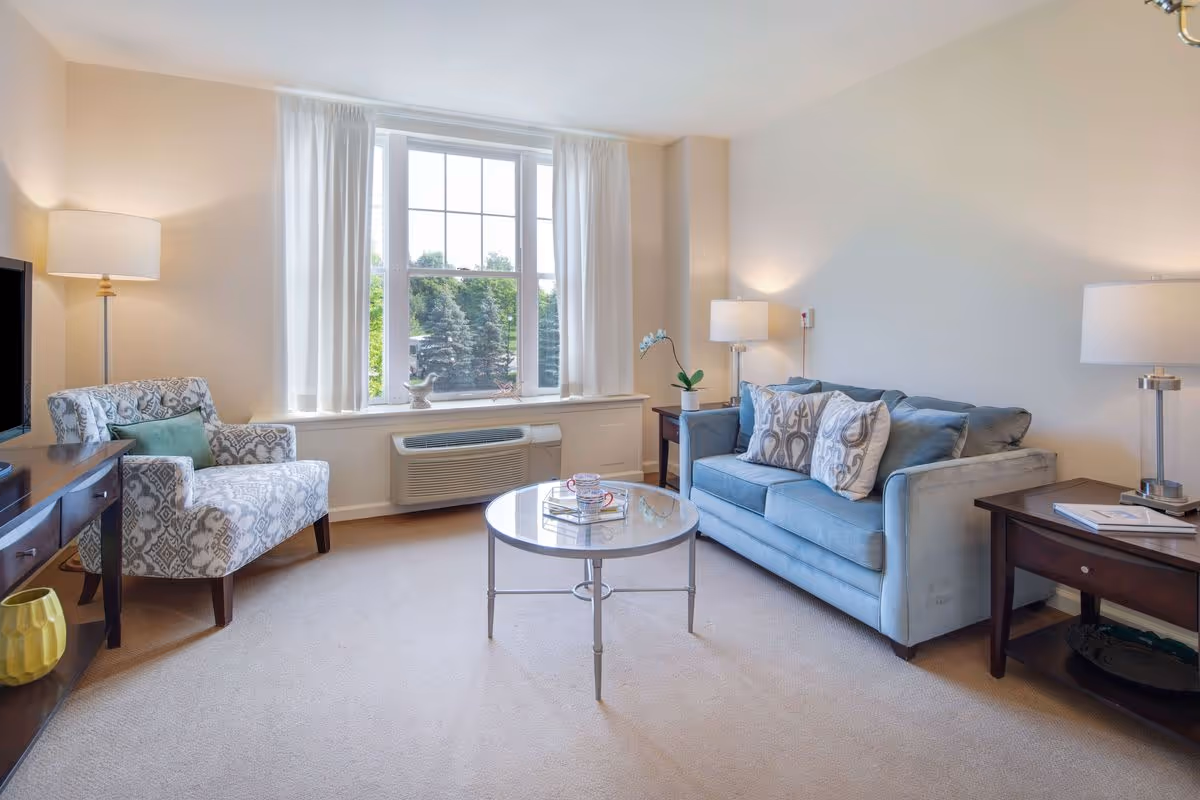 Bright living room with a blue sofa, patterned armchair, round glass coffee table, lamps, and a large window.