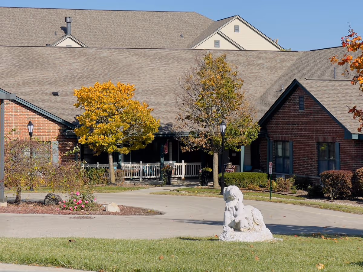 Front exterior of a brick senior living building with a porch, autumn trees, a driveway, and a small white statue on the lawn.