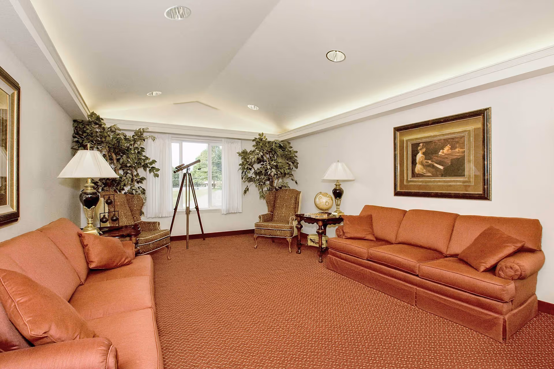 A cozy living room with two rust-colored sofas facing each other, two patterned armchairs, a telescope by a window with white curtains, two table lamps on wooden side tables, a framed painting on the wall, and two large potted plants in the corners.