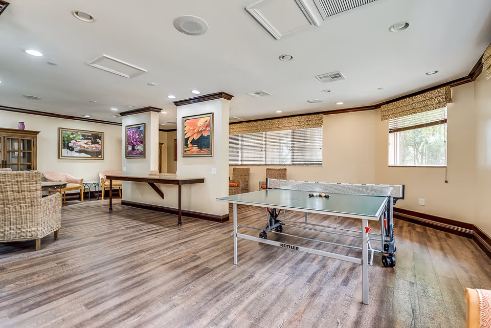 Bright senior living common room with a ping-pong table, seating area, and framed artwork on the walls.