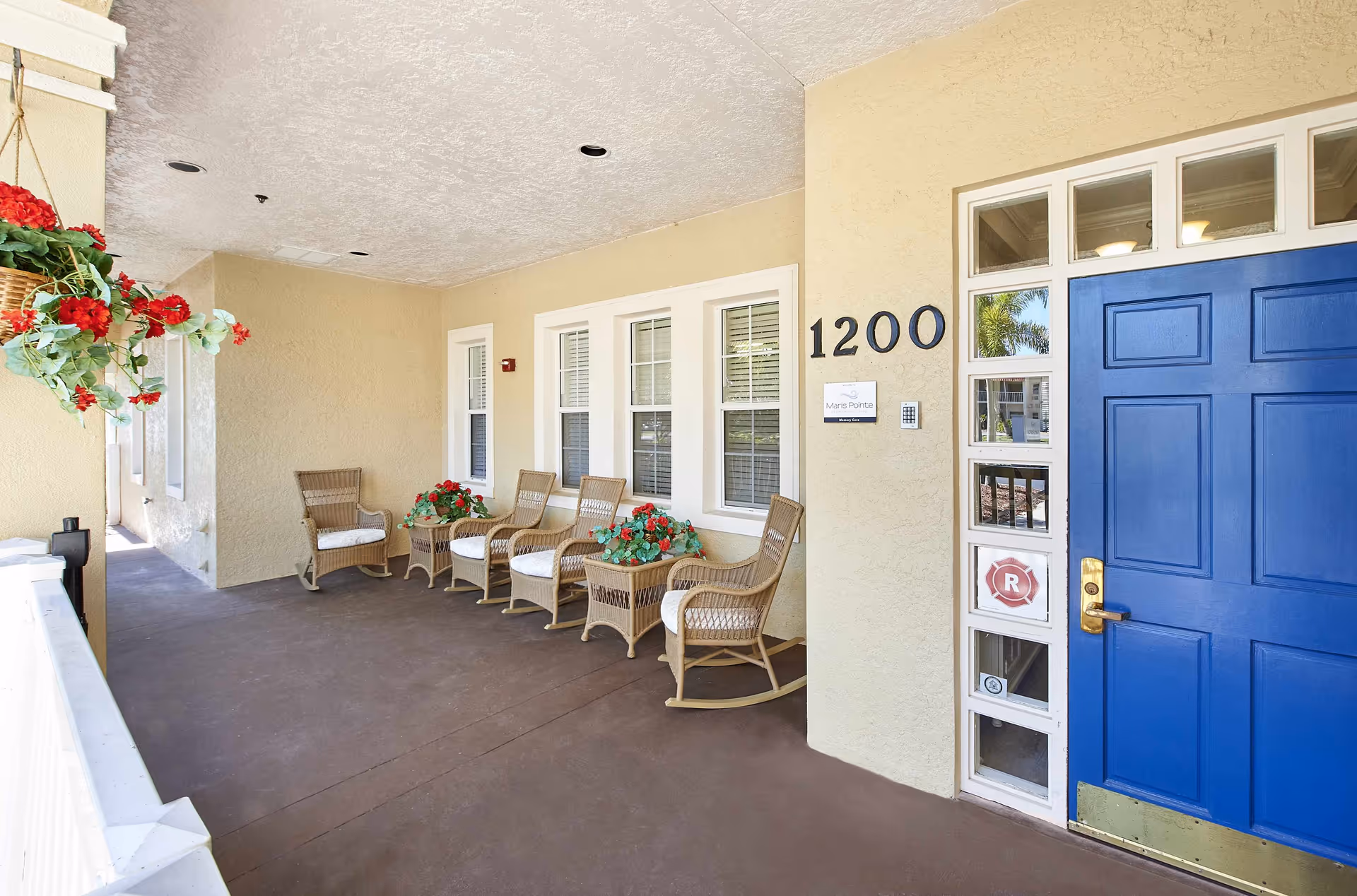 Covered entrance porch with a blue front door numbered 1200, wicker chairs and potted red flowers.