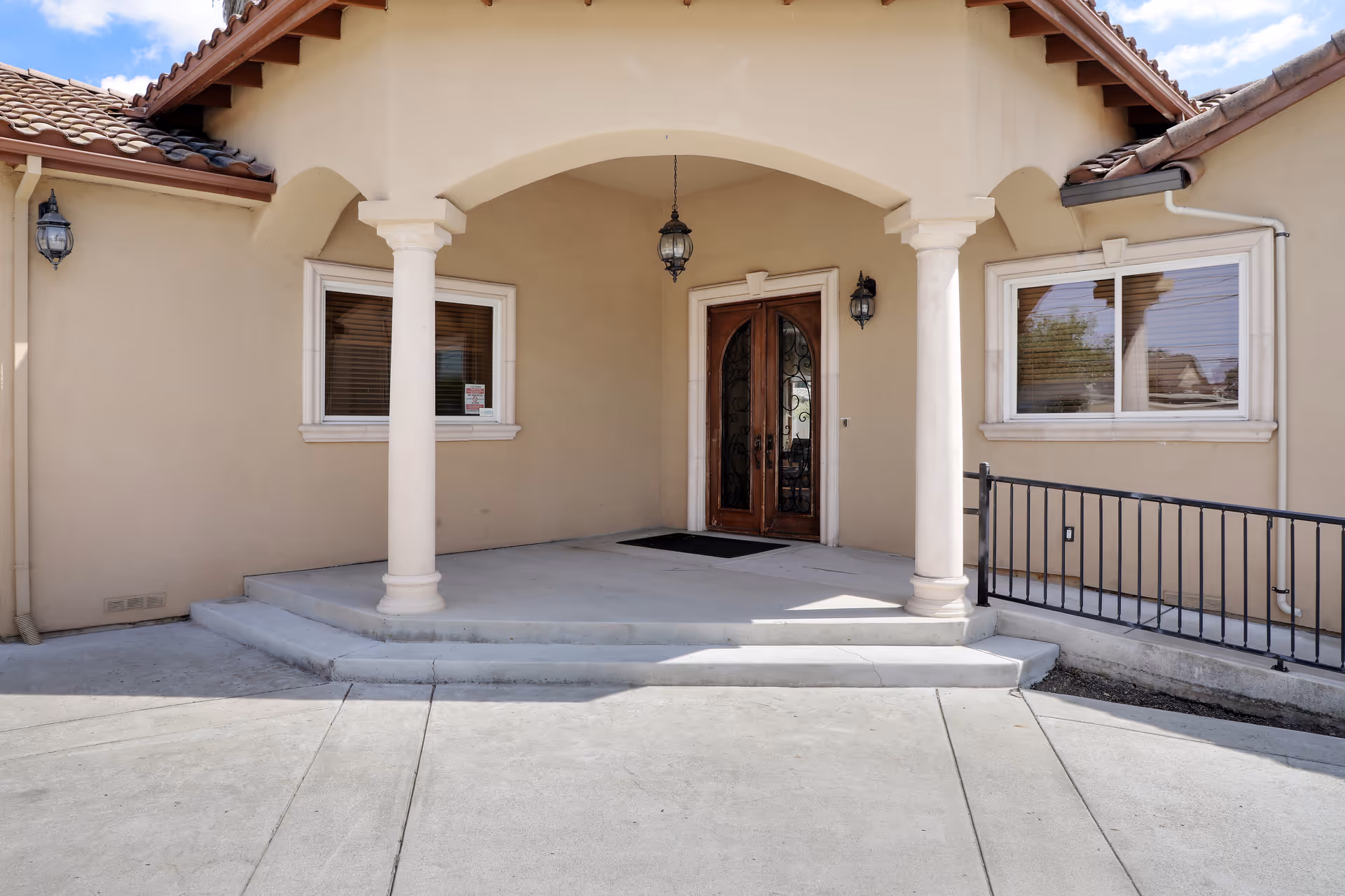 Front entrance of a building with a beige stucco exterior, two white columns supporting an arched porch roof, a double wooden door with decorative glass panels, two windows with white frames on either side, and a concrete ramp with a black metal railing leading up to the entrance.