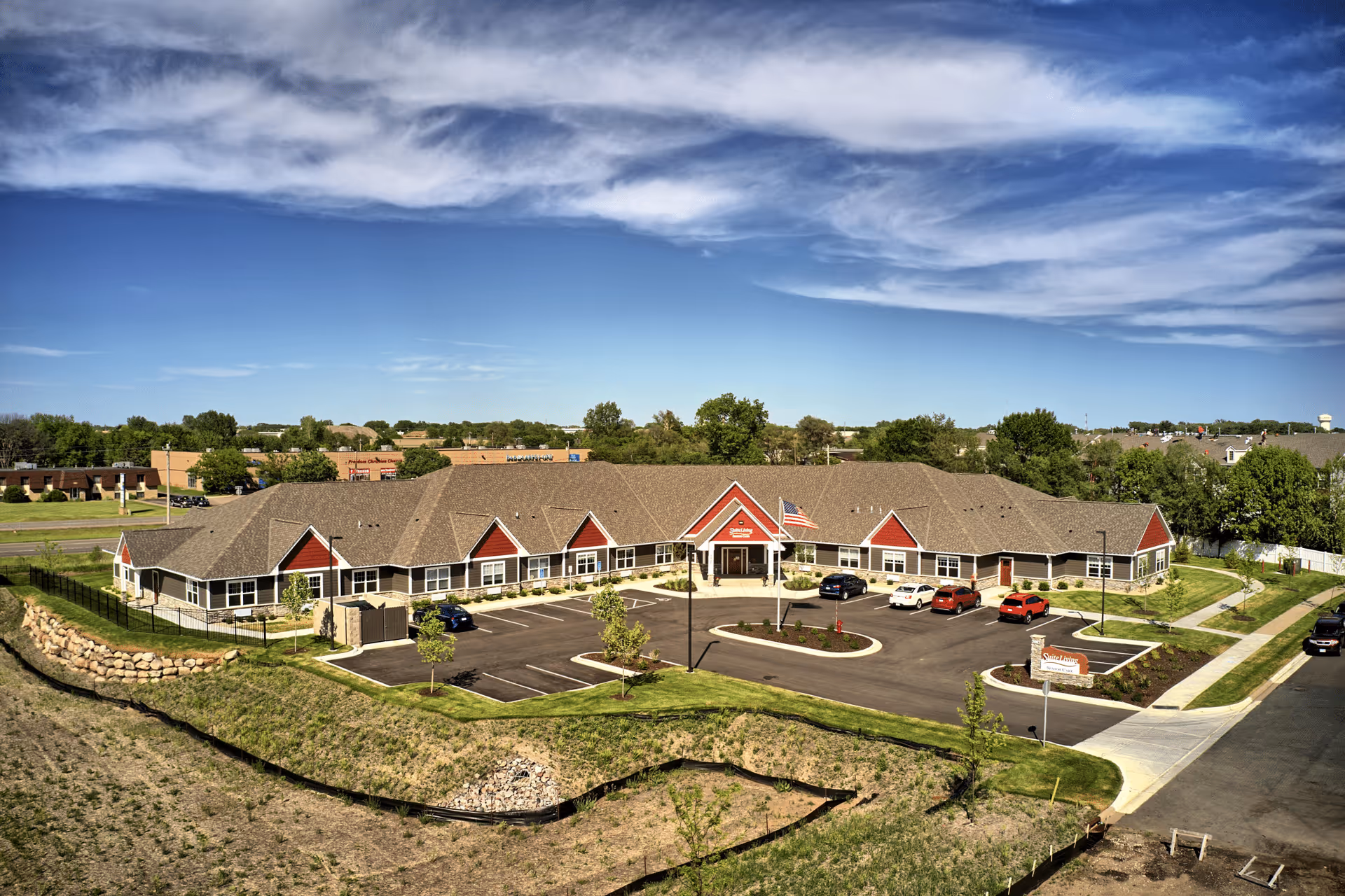 Aerial view of Suite Living of Brooklyn Park, a single-story senior care facility with a large parking lot, manicured lawns, and an American flag at the entrance. The building has a brown roof with red and white exterior accents, surrounded by trees and a clear blue sky with some clouds.