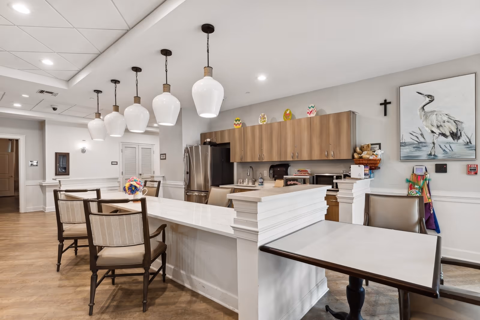 A modern kitchen and dining area in a senior living facility with a long white countertop island, several chairs, pendant lights hanging from the ceiling, wooden cabinets, stainless steel refrigerator, and a painting of a bird on the wall.