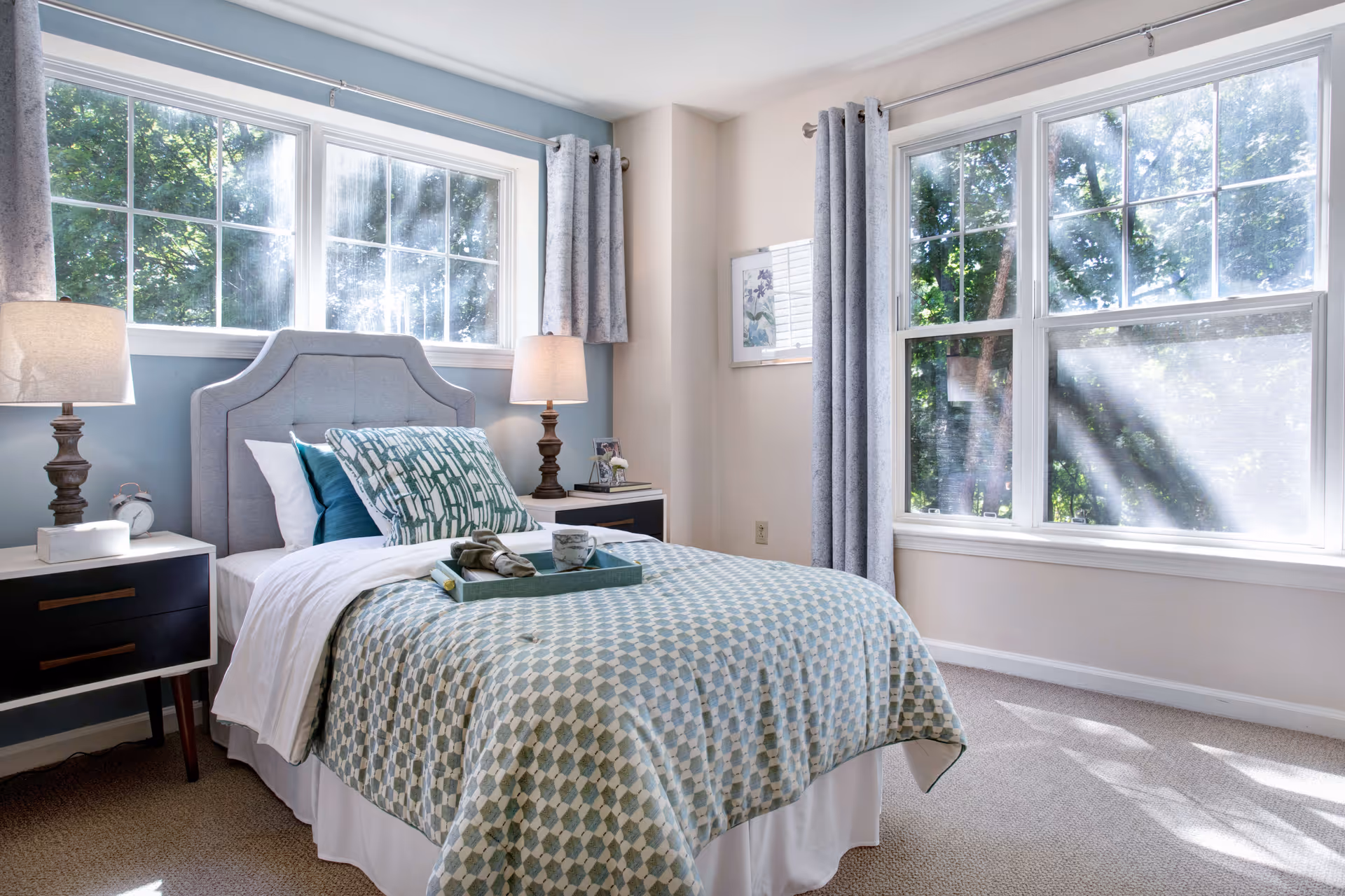 A bright and cozy bedroom with a single bed featuring a gray upholstered headboard, blue and white patterned bedding, and decorative pillows. There are two matching bedside tables with lamps on each side of the bed. Large windows with gray curtains allow natural light to fill the room, showing green trees outside.