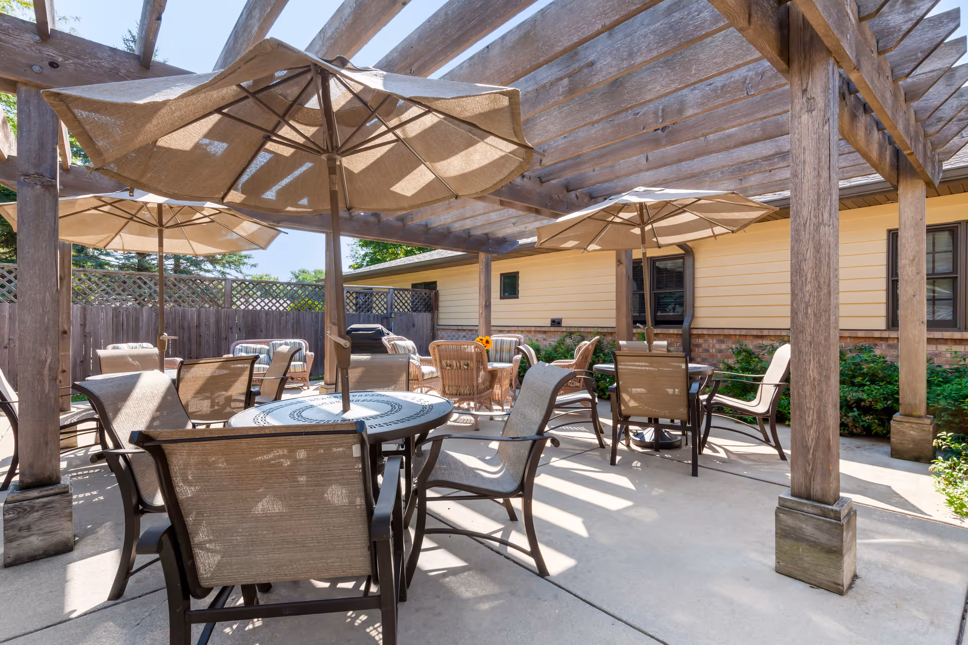 Outdoor patio area with multiple tables and chairs under large beige umbrellas and a wooden pergola. The patio is surrounded by a wooden fence and adjacent to a building with yellow siding and brick accents. Green shrubs are visible along the building.