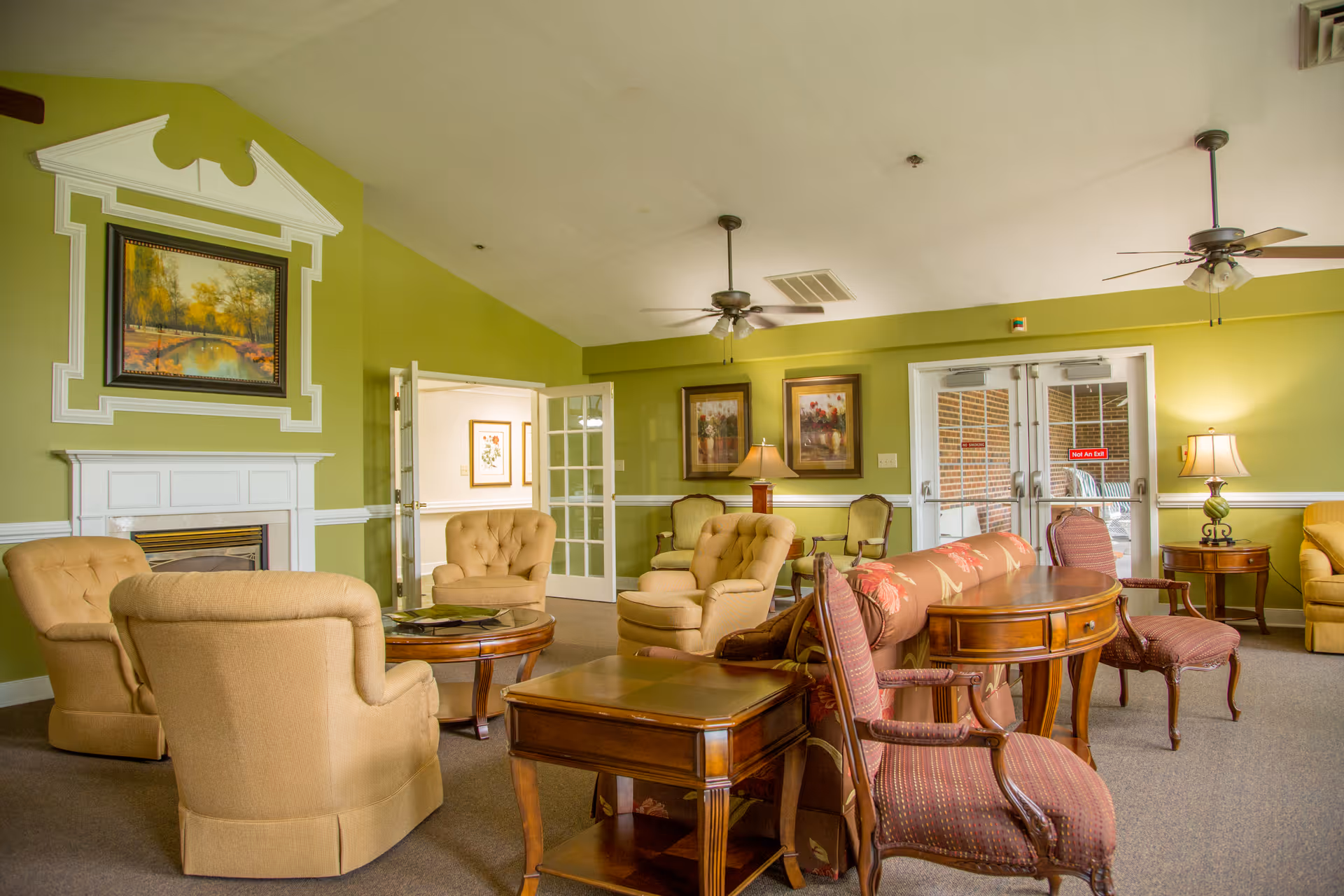 A cozy living room area with beige upholstered armchairs arranged around a wooden coffee table, a floral patterned sofa, and wooden side tables. The walls are painted light green with white trim, and there is a white fireplace with a framed landscape painting above it. Two ceiling fans and table lamps provide lighting, and glass double doors lead to an outdoor area.
