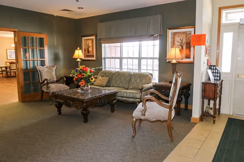 A cozy living room area in a senior living facility with a patterned sofa, two upholstered armchairs, a wooden coffee table with a floral arrangement, two table lamps, framed artwork on the walls, and a large window with a valance. The room has carpeted flooring and is adjacent to a tiled entryway with a door and a small table holding mail and a fire extinguisher.