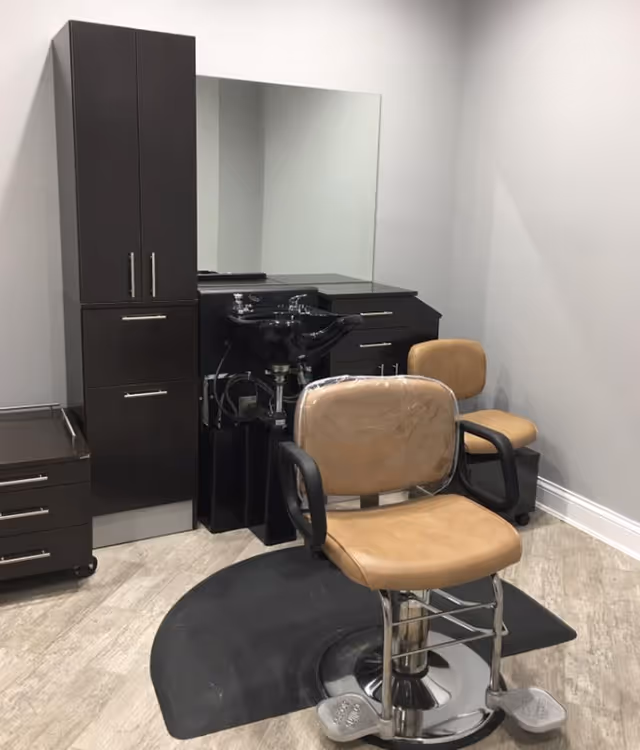 A salon styling station with a tan barber chair, shampoo sink, large mirror, and dark cabinetry in a clean room.