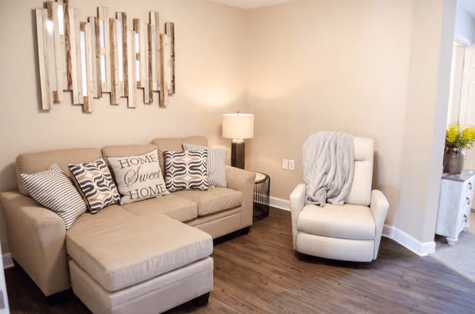 A cozy living room with a beige sectional sofa adorned with patterned pillows and a 'Home Sweet Home' pillow. Next to the sofa is a round side table with a lamp. A white armchair with a gray throw blanket is positioned nearby. The room has wooden flooring and neutral-colored walls with a decorative wall mirror above the sofa.