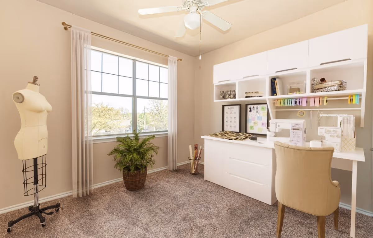 A bright sewing or craft room with a large window letting in natural light. The room features a white desk with sewing machines, colorful ribbons hanging above, and framed artwork on the desk. A beige upholstered chair is positioned at the desk. To the left, there is a dress form mannequin and a potted plant on the carpeted floor. The walls are light-colored, and a ceiling fan with a light fixture is visible.