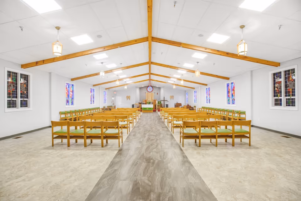 Interior view of a chapel or worship room with wooden beams on the ceiling, stained glass windows on both sides, rows of wooden chairs with green cushions arranged facing an altar at the far end, and pendant lights hanging from the ceiling.