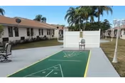 Outdoor recreational area at Sunny Hills Assisted Living Facility Homestead featuring a shuffleboard court with a chair placed at the far end. The area is surrounded by single-story buildings with palm trees in the background and a paved walkway.